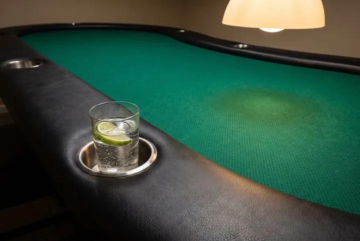 Close view of a poker table edge: a black padded rail with a glass of sparkling water and lime in its cup holder, empty green felt beyond.